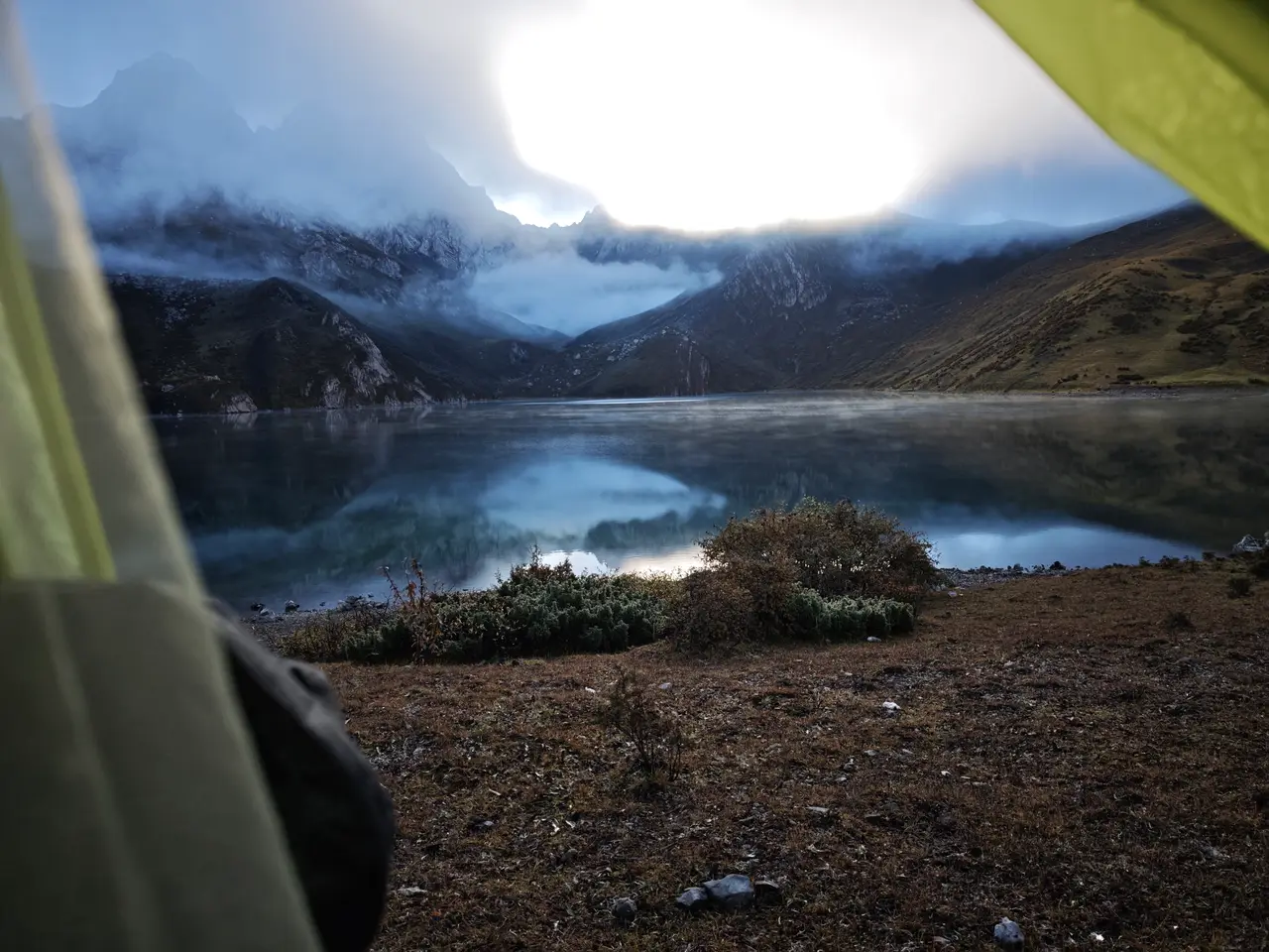 帐篷边的湖面在逆光里发亮，清晨云层和山影一起压在水边。